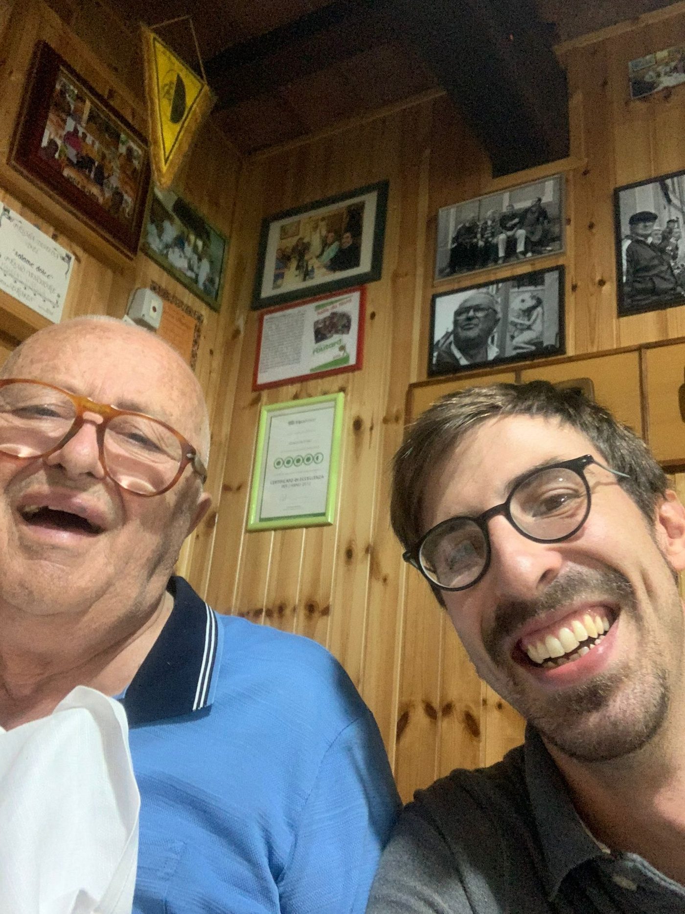 Alessandro Dolcini ed Ermes, sorridenti fianco a fianco nella sala della trattoria, sotto la parete di fotografie incorniciate