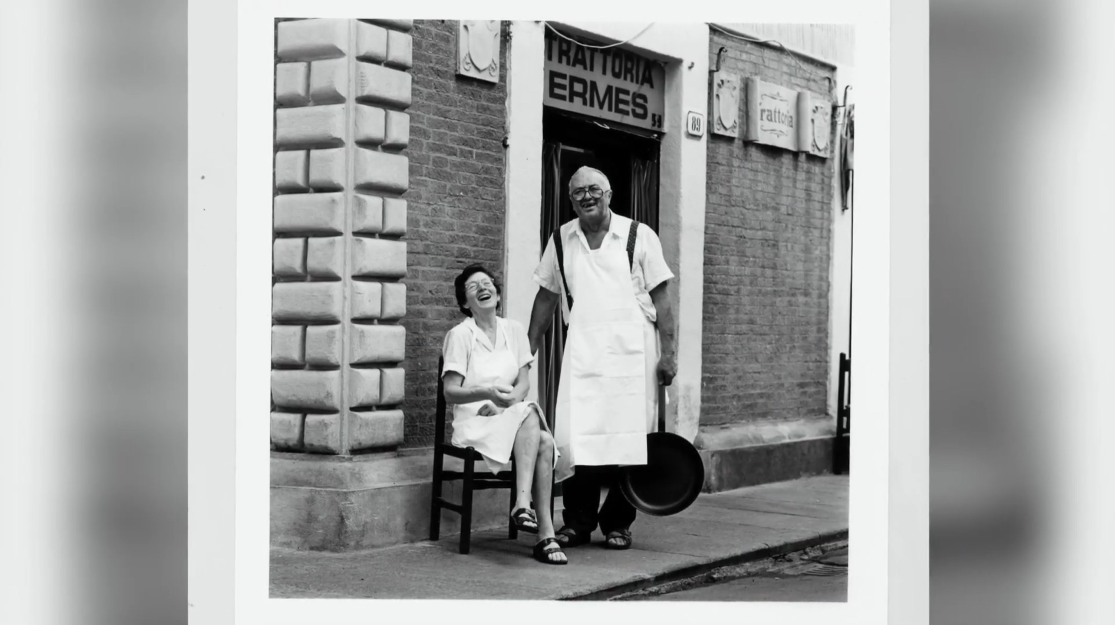 Ermes, nel suo grembiule bianco e con una padella in mano, e Bruna, seduta su una sedia, davanti all'ingresso della Trattoria Ermes a Modena negli anni d'oro